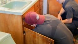 Two people in work uniforms kneel by an open bathroom cabinet under a sink, inspecting or repairing plumbing, possibly addressing repeated drain clogs.