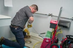 A plumber kneels on the floor while using a drain cleaning machine to unclog a pipe in a bathroom, addressing repeated drain clogs with expert care.
